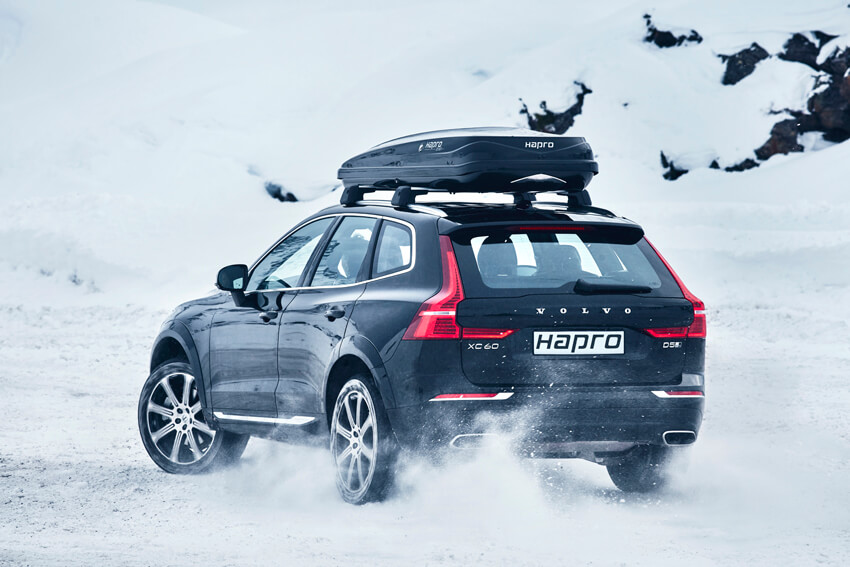 A black 2022 Volvo XC60 with a Hapro roof box drives on a snowy road surrounded by snowy rocks. The car is in motion, kicking up snow from the ground.