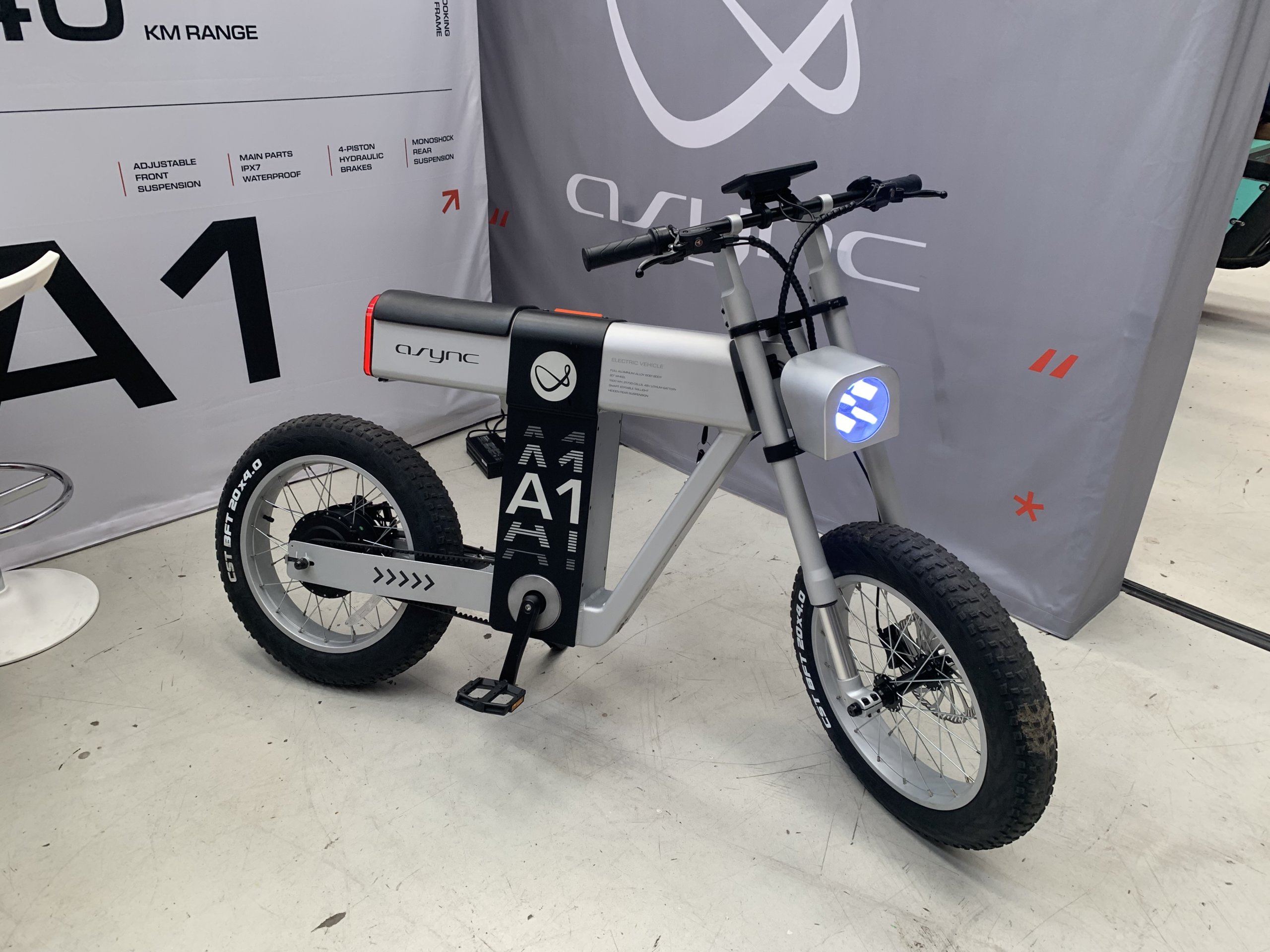 Exhibition display of a sleek, modern A1 electric bike by Qync, featuring chunky off-road tires, a prominent frame-mounted battery, and a front LED light, set against a backdrop with specifications listed such as 'KM Range' and 'Adjustable Front Suspension'.
