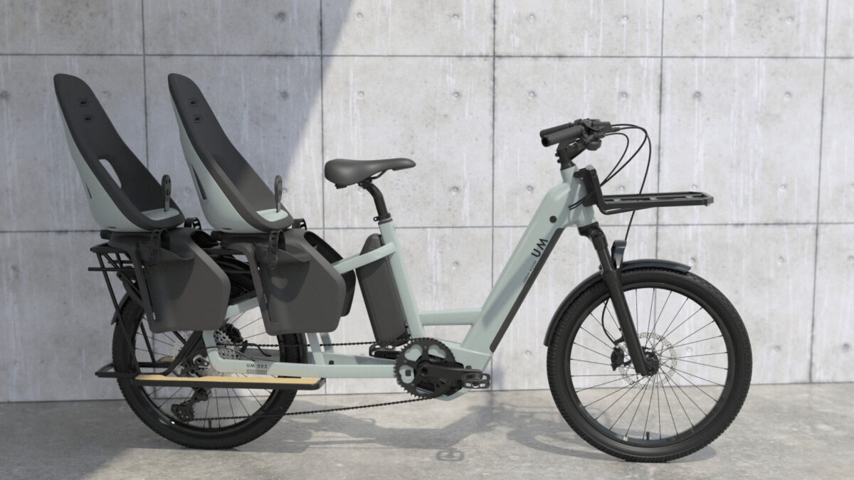A modern electric cargo bike, perfect for urban environments, boasts a sleek gray frame parked against a concrete wall. It features two black child seats on the rear rack, each equipped with footrests and safety harnesses. The front of this urban mover has a rack for carrying additional cargo.